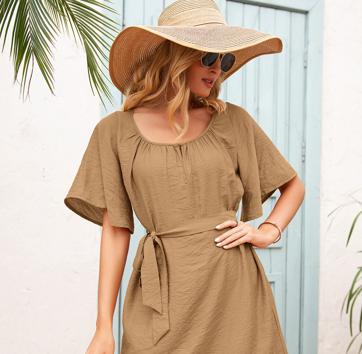 Woman wearing a brown dress and wide-brimmed hat with sunglasses, standing against a light blue wall.